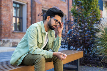 Sick man sitting on bench outside office building, Indian man having severe headache and dizziness migraine, overworked and overworked worker in casual clothes