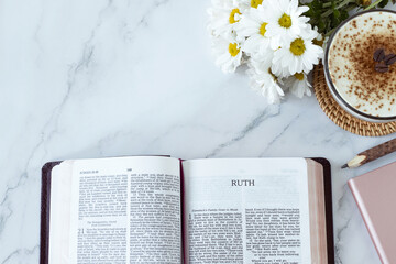 Ruth open bible book with coffee cup, flowers, pencil, and notebook on white marble background. Top...
