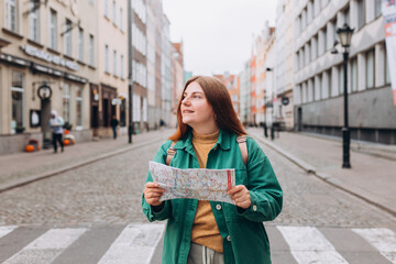 Portrait redhead woman with paper map on urban street. Happy tourist travels in Europe. Vacation concept by exploring interesting places to travel. 30s Woman Searching locations at autumn day, Gdansk