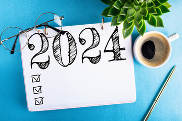 New year resolutions 2024 on desk. 2024 goals list with notebook, coffee cup, plant on blue table....