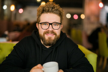 Bearded curly man smiling confident drinking coffee in restaurant or coffee shop. Millennial...