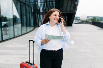 Beautiful redhead woman holding paper map and talking on smartphone outdoors. Lady with suitcase choosing route via paper map, navigating her urban journey