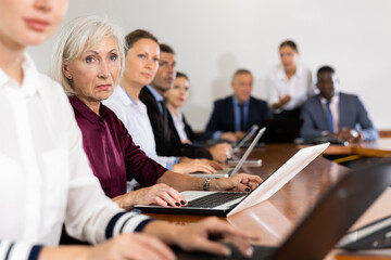 Portrait of elegant elderly white gray-haired female manager attending business meeting in conference room and watching colleague's presentation together with coworkers