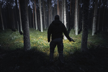 Silhouette of a man with a pistol and a flashlight in a dark forest, viewed from the back. - obrazy, fototapety, plakaty