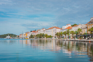 Promenade with palms and other trees and houses in the background in the city of Mali Losinj, on croatian island of Losinj, on a sunny day in autumn. Almost no people visible. - obrazy, fototapety, plakaty