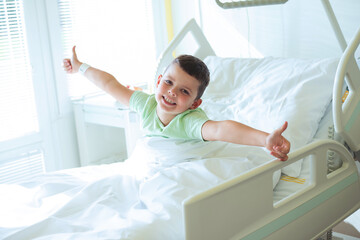 Fototapeta na wymiar Portrait of a smiling European seven year old boy in a light green T-shirt lying on the bed in a hospital room and showing thumbs up