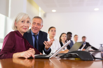 Obraz premium Positive senior woman manager sitting at desk beside colleagues and using laptop during conference.