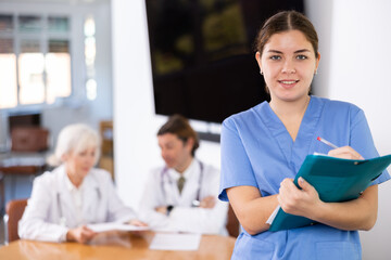 Portrait of smiling young female doctor in blue uniform meeting patient in medical office, filling out medical form at clipboard
