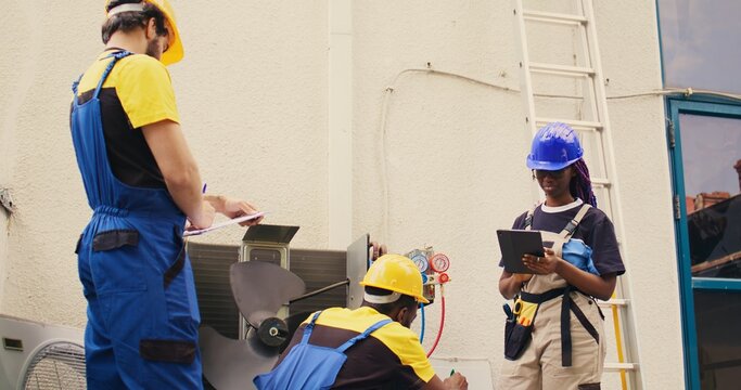 Trained diverse teamworking servicemen cleaning external condenser internal coolants, checking freon level and calibrating thermostats while coworker checks maintenance procedures on tablet