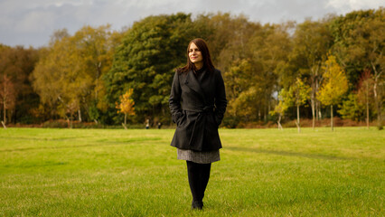 Full length shot of a middle-aged woman with medium length brown hair, walking in a park in the autumn with trees behind. - Powered by Adobe