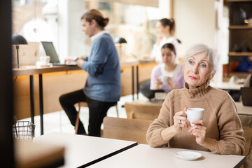 Sad elderly woman drinking coffee or tea at the bar counter alone