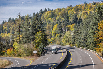 Autumn landscaping with forest on the hill and middle duty rig semi truck with box trailer running on the turning highway road with entrance fork intersection