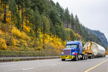 Blue big rig bonnet semi truck tractor with oversize load sign transporting part of oversized wind turbine pillar running on the picturesque autumn road in Columbia Gorge