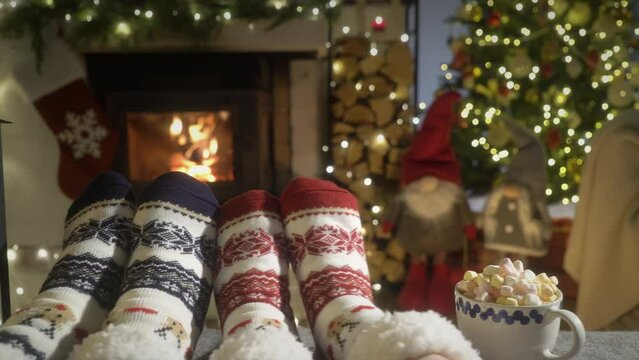 Couple enjoying cocoa with marshmallows by warm fireplace and warming up their feet in woolen socks with Christmas ornaments and Christmas tree and decorations background. Cozy Winter, Christmas and