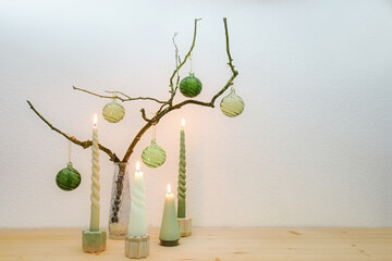 Advent and Christmas decoration with four different candles and green glass balls on a bare branch in a vase on a wooden sideboard against a light wall, copy space