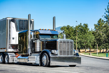 Powerful black big rig long hauler classic semi truck tractor with loaded refrigerator semi trailer standing on the highway road rest area parking lot