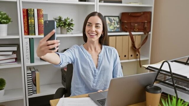 Radiant young hispanic woman captures selfie with smartphone during office work, a snapshot into her successful business lifestyle at the elegant workplace