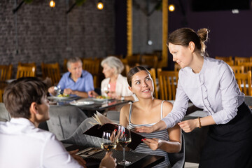 Young male and female colleagues having nice time in sparsely populated cozy restaurant. Waitress answers questions of visitors, helps to choose diet dish for late dinner in menu - obrazy, fototapety, plakaty