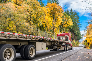 Red big rig semi truck with empty flat bed railer running on the narrow road with yellow autumn trees on the sides