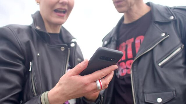 Woman holds phone close-up as a couple engages in animated discussion while looking at smartphone screen. They are actively sharing and discussing content, possibly photos, messages, or information.