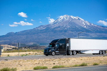Stylish black big rig bonnet semi truck tractor transporting cargo in refrigerator semi trailer running on the highway road past the snowy mountain in California