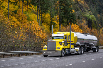 Yellow bright classic big rig semi truck tractor transporting liquid cargo in tank semi trailer running on the picturesque autumn road with rock mountain on the side