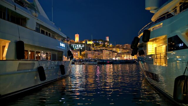 View of the sea port of Cannes with moored yachts at night, France. Cityscape with Castre museum on a hill in the distance, illumination