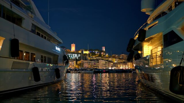 View of the sea port of Cannes with moored yachts at night, France. Cityscape with Castre museum on a hill in the distance, illumination
