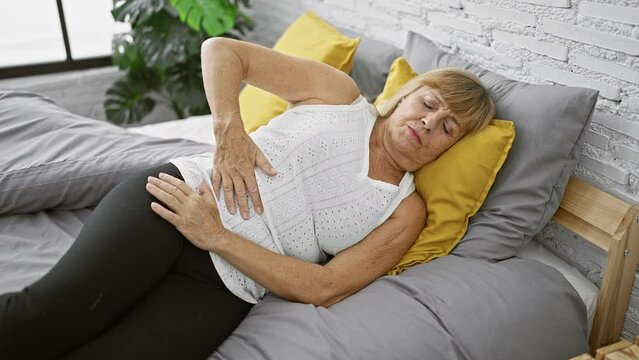 Middle age blonde woman suffering immense stomach pain, touching her abdomen unhappily while lying in bed, illustrating the struggle of indoor illness.