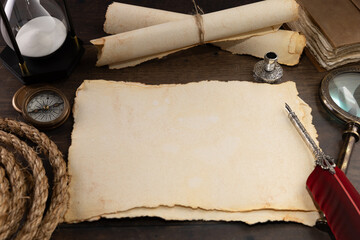 Rustic old sheet of parchment in landscape orientation with room for copy on a wooden surface with ink quill and rolled parchments nearby.
