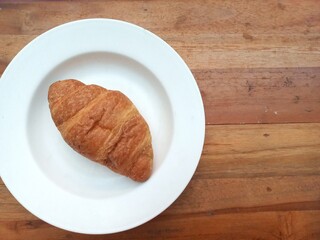 Top view photo of croissant on white plate on wooden table. This bread has a layered texture, a high level of patience is required to make this cake. Flay lay. Close up. Space for text.
