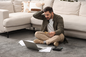 Confused man with laptop, debt notifications and credit card planning budget at home. Financial...