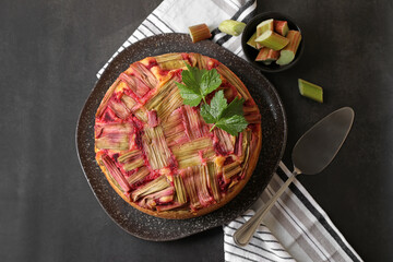 Freshly baked rhubarb pie, cut stalks and cake server on black table, flat lay