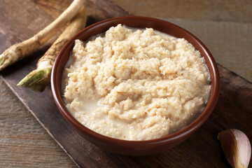 Spicy horseradish sauce in bowl, roots and garlic on wooden table, closeup