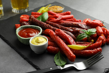 Different thin dry smoked sausages, basil and sauces on grey table, closeup