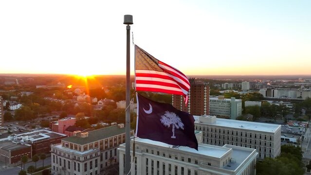 American and South Carolina flags waving atop capitol building in downtown Columbia, SC. Aerial orbit shot with government buildings during bright sunrise.