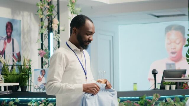 Employee restocking luxury fashion boutique elegant clothes on racks. African american worker adding high class men shirts and other garments on hangers in trendy clothing store