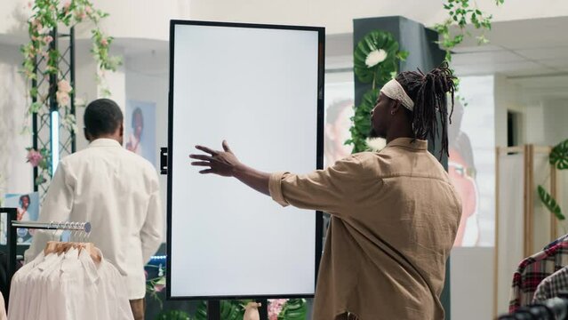 Shopper in second hand clothing shop choosing between clothes using isolated digital screen. Buyer looking at new collection of garments in discount store using augmented reality kiosk
