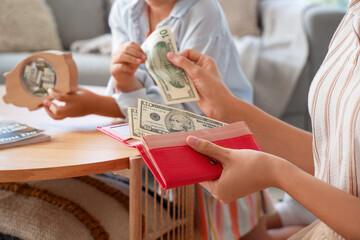 Little girl with her mother putting money into piggy bank at home, closeup
