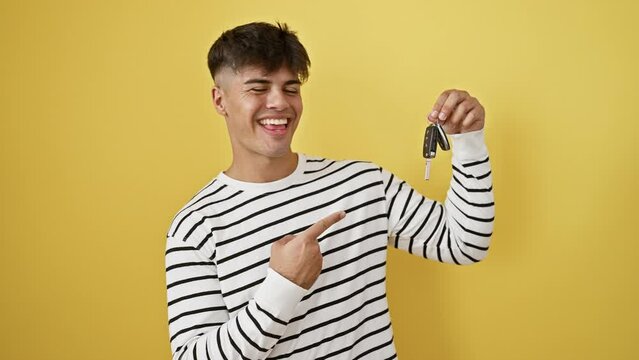 Cheerful young hispanic man, confidently pointing with a key car in hand. portrait of his happy expression, marking joy and emotion isolated on a radiant yellow background.