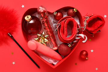 Box in shape of heart with different sex toys and Christmas decor on red background