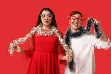 Young couple in decorative eyeglasses with Christmas tinsel on red background