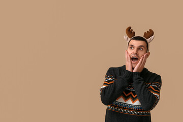 Shocked young man in reindeer horns on beige background