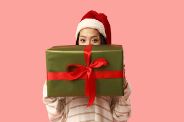 Young Asian woman in Santa hat with Christmas gift box on pink background