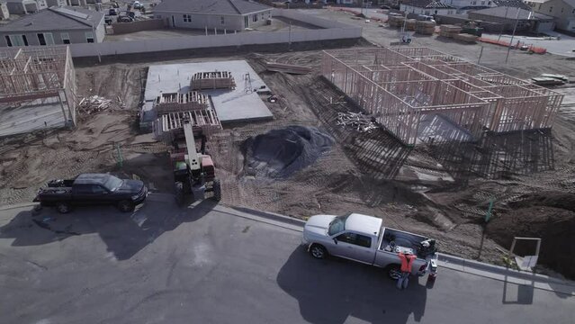 A drone observes an extendable forklift in action, gently lowering the third crate of wood onto a cement foundation within a new neighborhood construction site where no walls have been erected yet