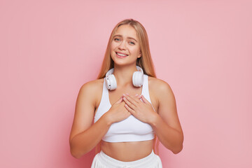Atractive blonde woman in white top with white headphones on her neck stands on pink studio backdrop pressing hands to her heart, beauty concept, copy space