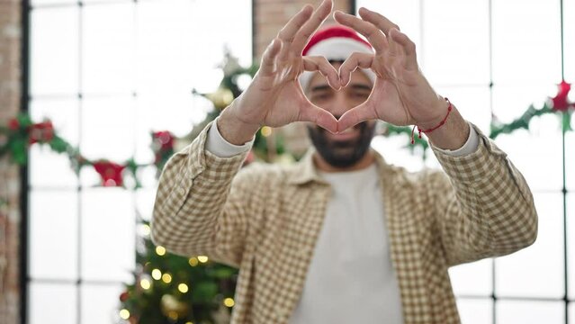 Young hispanic man doing heart gesture celebrating christmas at home