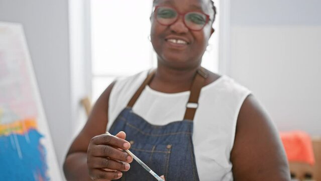 Cheerful african american woman artist, confidently holding paintbrush and palette, smiling bright in indoor art studio