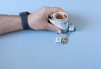 A man's hand holding a key and a roll of money, on a blue background. Concept - buying a house.