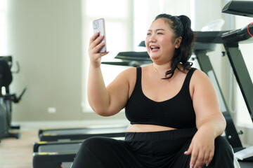 Fototapeta premium Chubby woman sits and rests after exercising in the fitness center. Video call with friends via smartphone To invite you to exercise. Happiness of a cute Asian overweight woman trying to lose weight